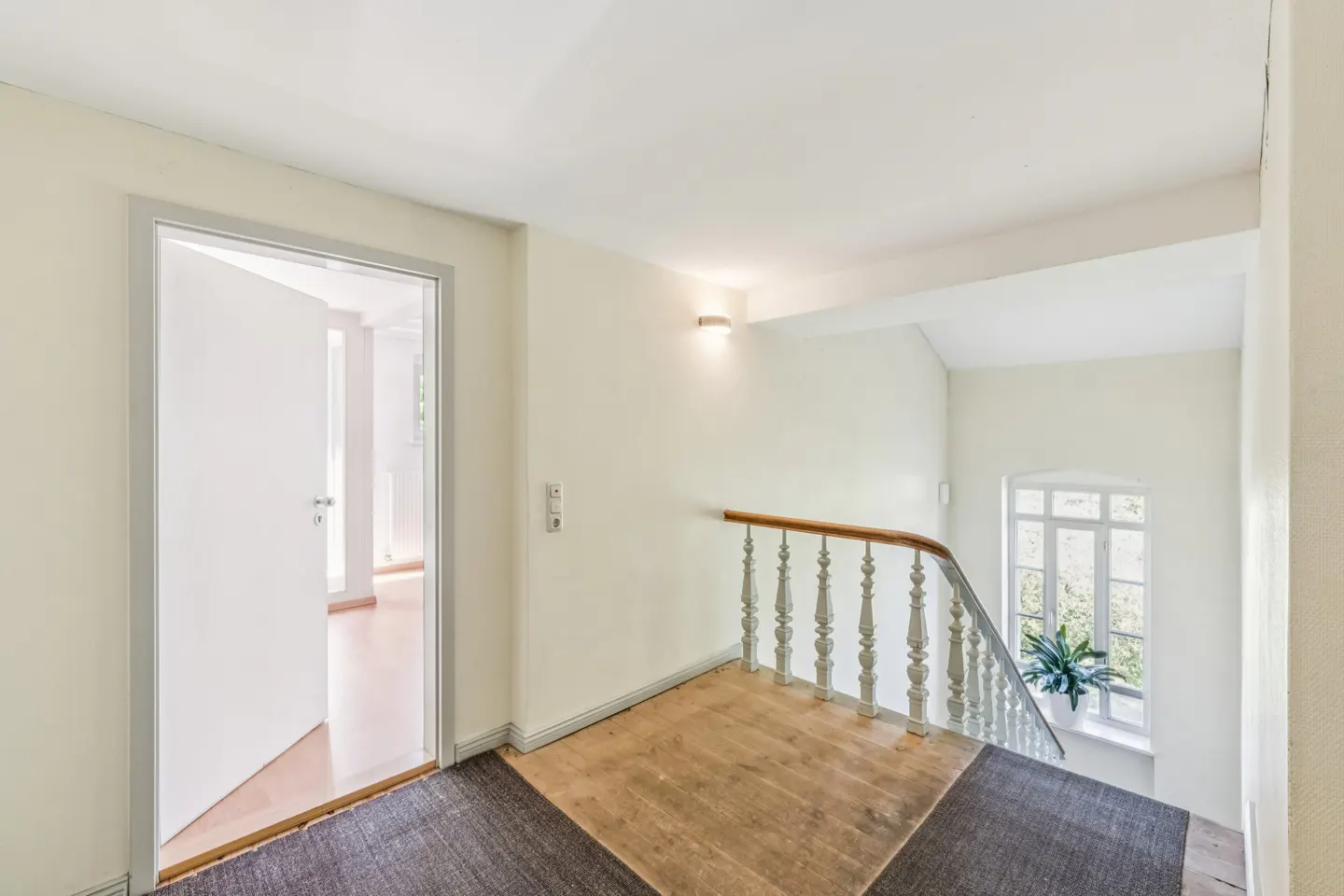 Bright hallway with wooden floors, a white door leading to a room, and a staircase with a wooden railing and window.