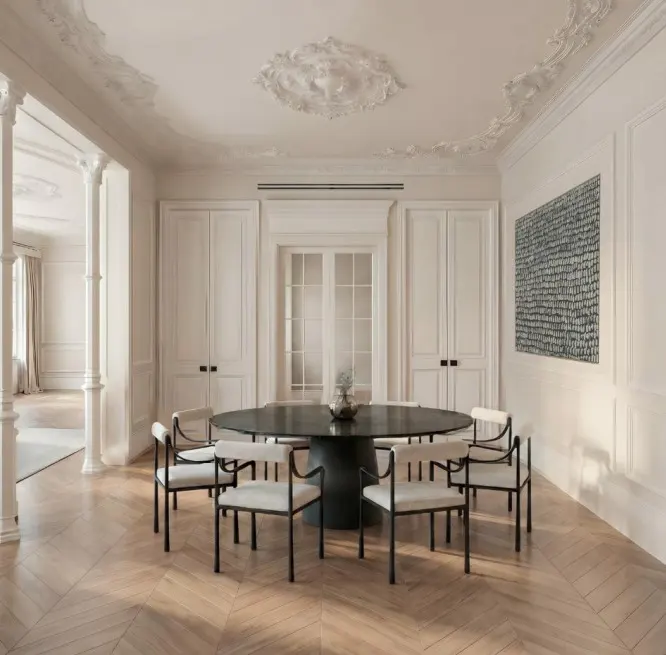 Bright dining room with herringbone wood floors, white walls, and ornate ceiling. A black round table is surrounded by six white chairs.