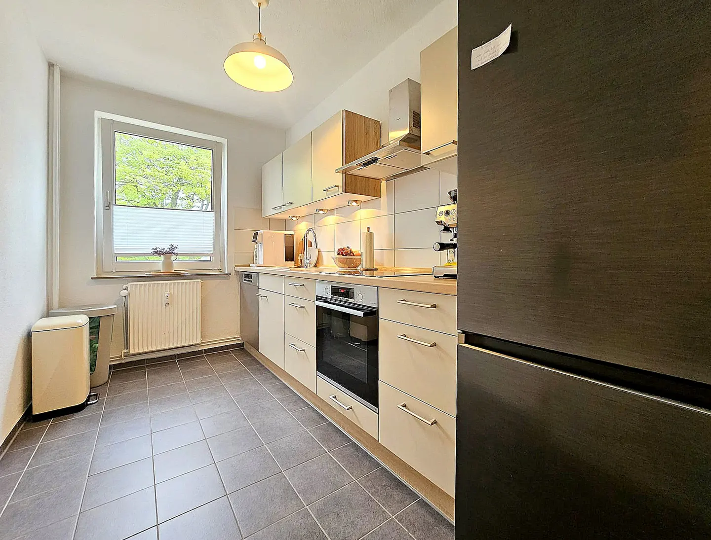 Bright kitchen with gray tile floor, beige cabinets, and a black refrigerator. A window with blinds is on the left.