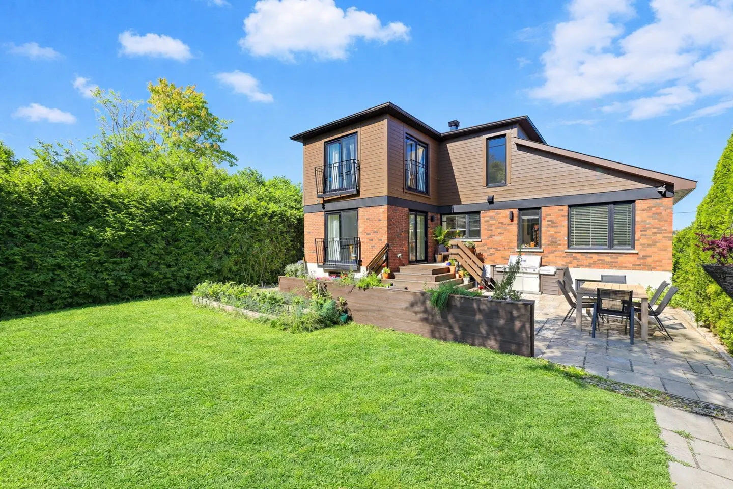 Two-story brick and brown siding home with a backyard patio, dining set, and lush green lawn under a blue sky.