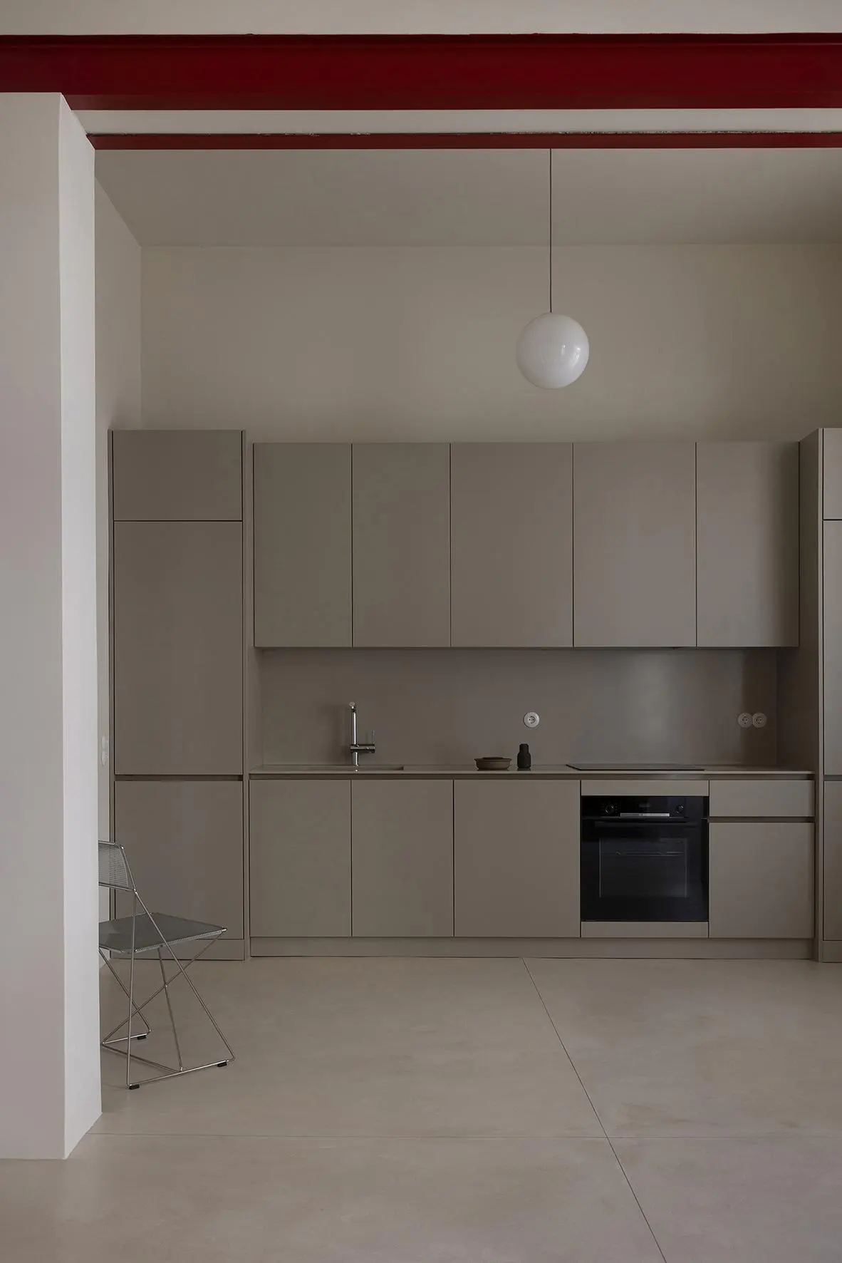 Minimalist kitchen with gray cabinets, stainless steel sink, and black oven. A white pendant light hangs above, and red beams accent the ceiling.