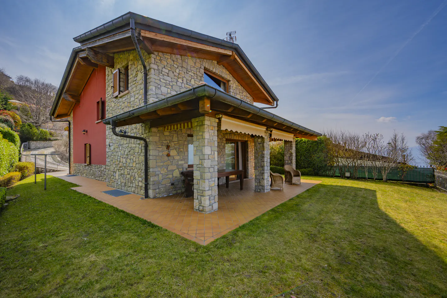 Stone house with red accent wall, covered patio with furniture, and green lawn under a blue sky.