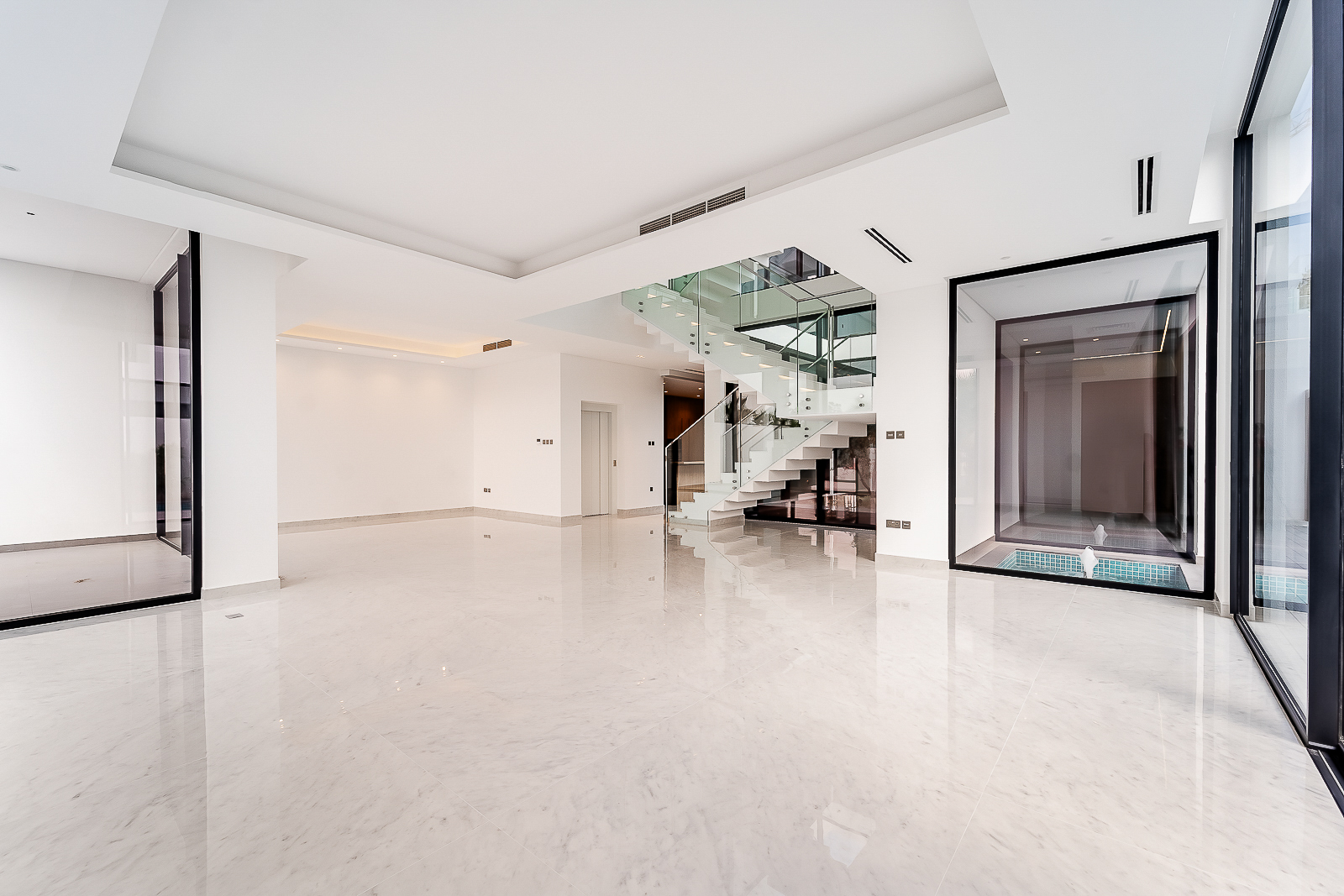 Modern, minimalist living room with white marble floors, a glass-railed staircase, and large windows overlooking a courtyard. [AI-generated text]