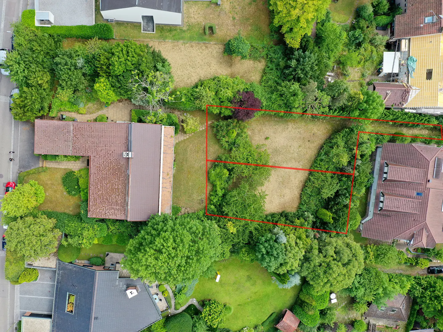 Aerial view of a residential property with a vacant plot outlined in red, surrounded by lush green trees and houses.