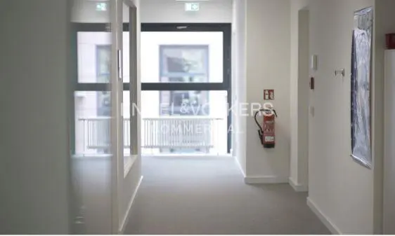 Hallway with gray carpet, white walls, and a fire extinguisher. A window at the end shows a building exterior.