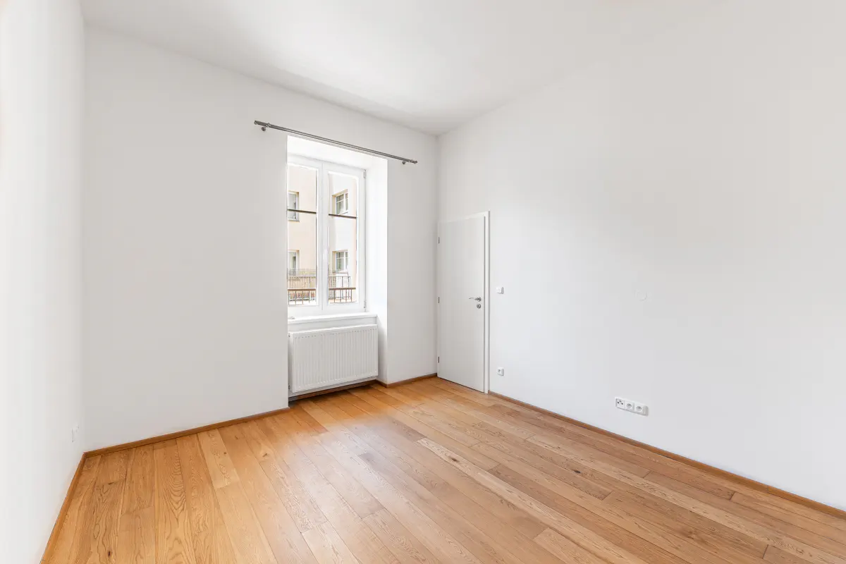Bright, empty room with hardwood floors, white walls, a window with a radiator, and a closed white door.