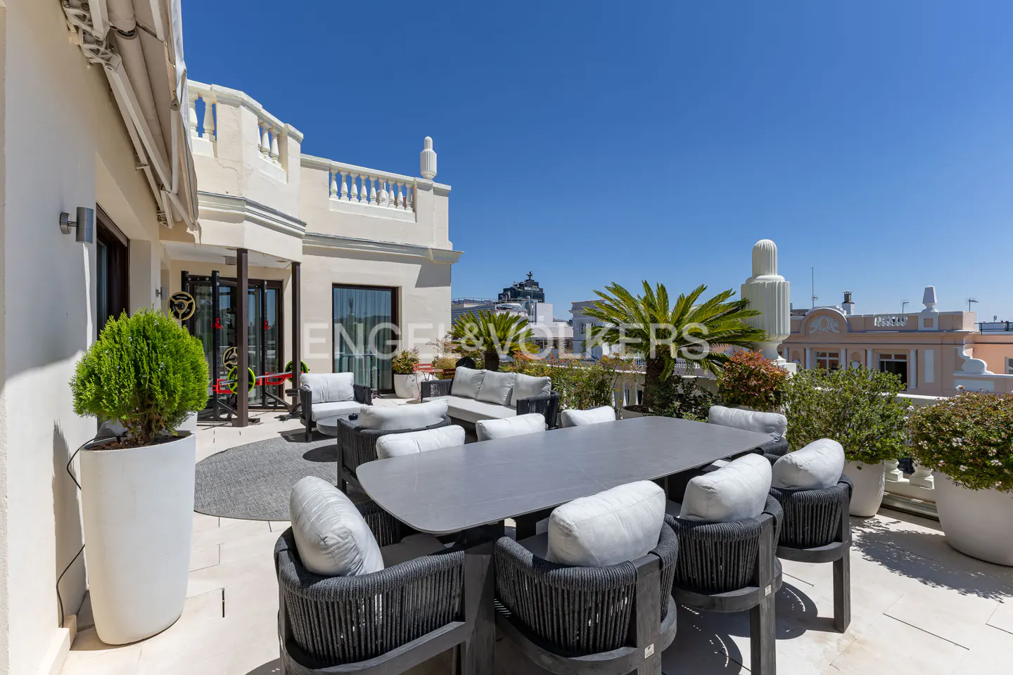 Outdoor rooftop patio with a large gray table, chairs, and white cushions. Plants and city views in the background.