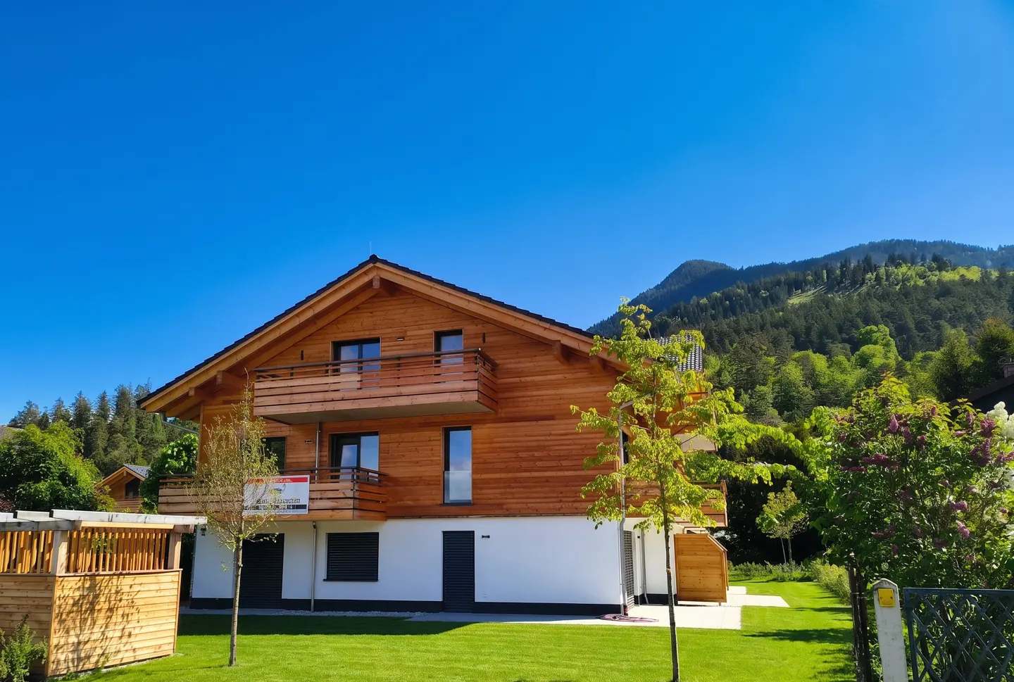 Two-story wood house with a balcony, white foundation, green lawn, and mountain backdrop under a clear blue sky.