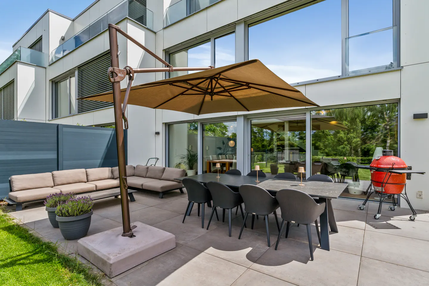 Modern patio with a tan cantilever umbrella, beige sectional sofa, dining set with gray chairs, and a red charcoal grill.