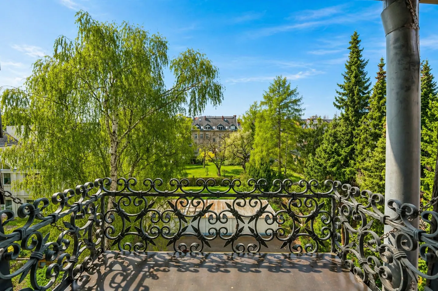 View from a balcony with ornate black iron railings overlooking green trees and a building under a blue sky.