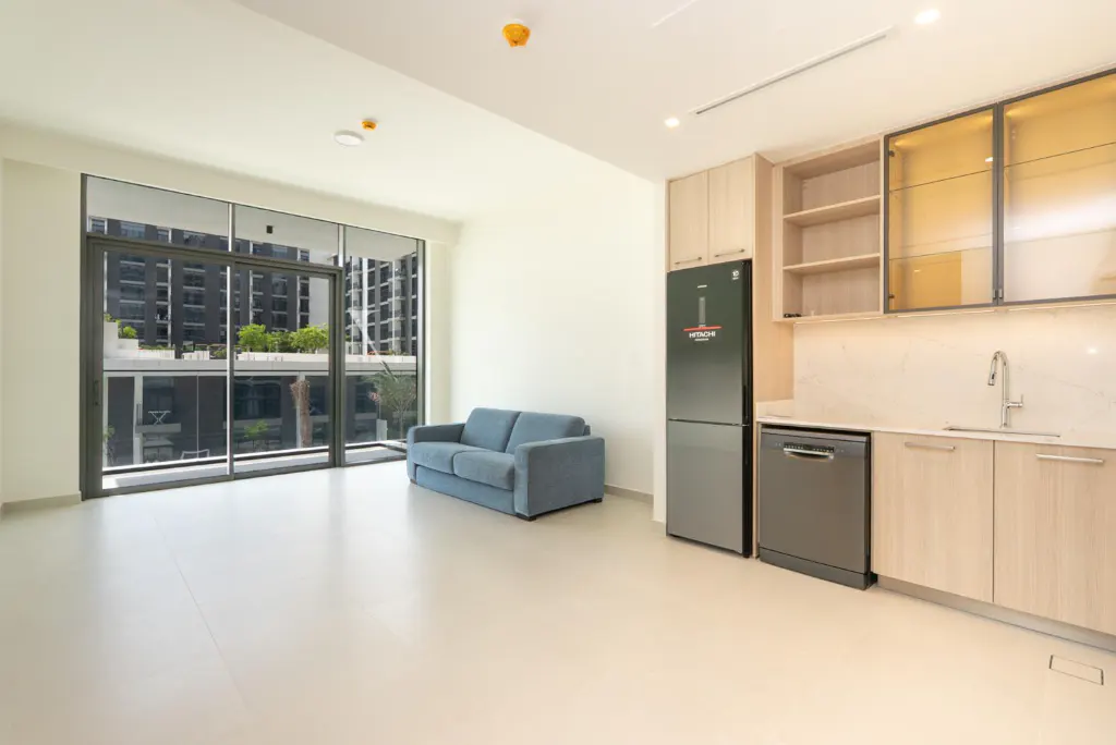 Bright, modern apartment with a blue sofa near a large window. A kitchen area features light wood cabinets and stainless steel appliances.