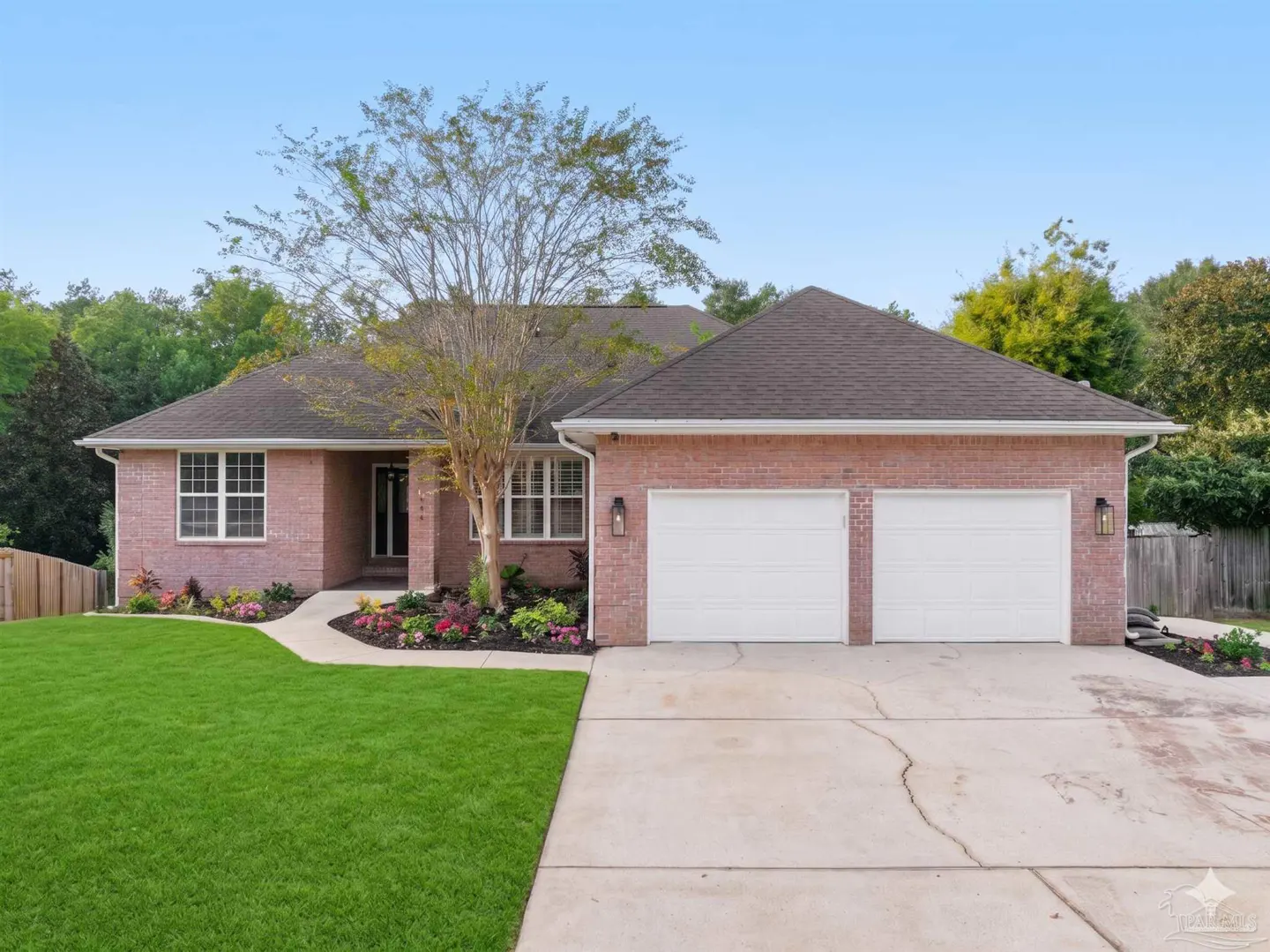 A single-story red brick house with a brown roof, white garage doors, green lawn, and blue sky.