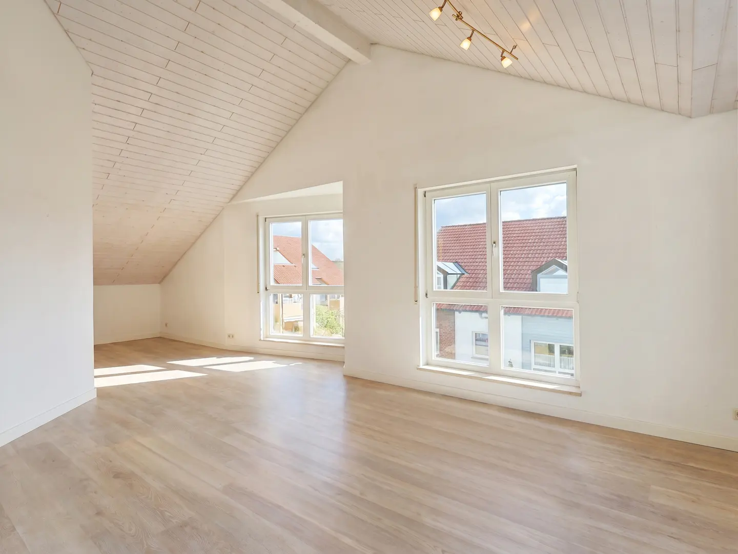 Bright, empty attic room with white walls, wood floors, and a white plank ceiling. Two windows show houses outside.