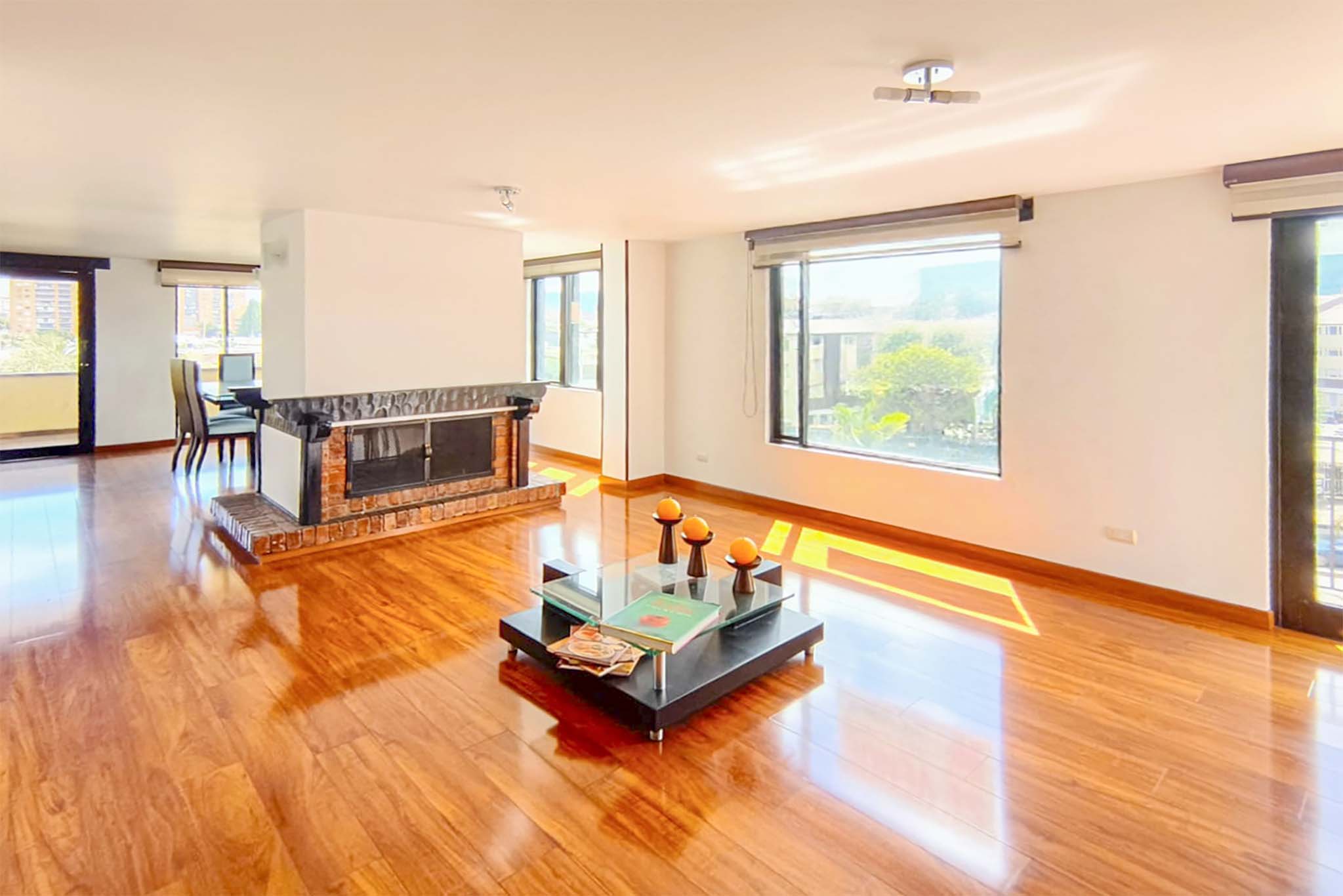 Bright living room with hardwood floors, a brick fireplace, and large windows. A glass coffee table sits in the center of the room.