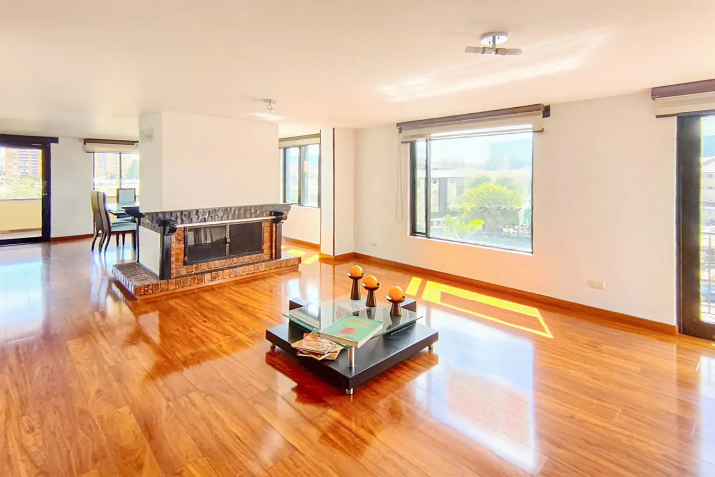 Bright living room with hardwood floors, a brick fireplace, and large windows. A glass coffee table sits in the center of the room.