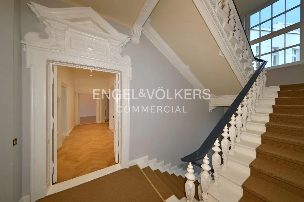 Interior view of a staircase with brown carpet, white balusters, and a dark blue handrail. A doorway leads to a room with herringbone wood floors.