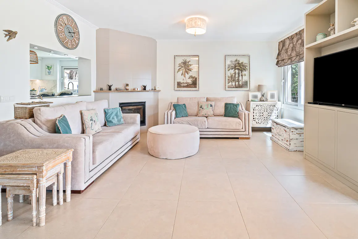 Living room with beige sofas, a fireplace, and palm tree art. A large clock hangs above the sofa, and a TV is on the right.