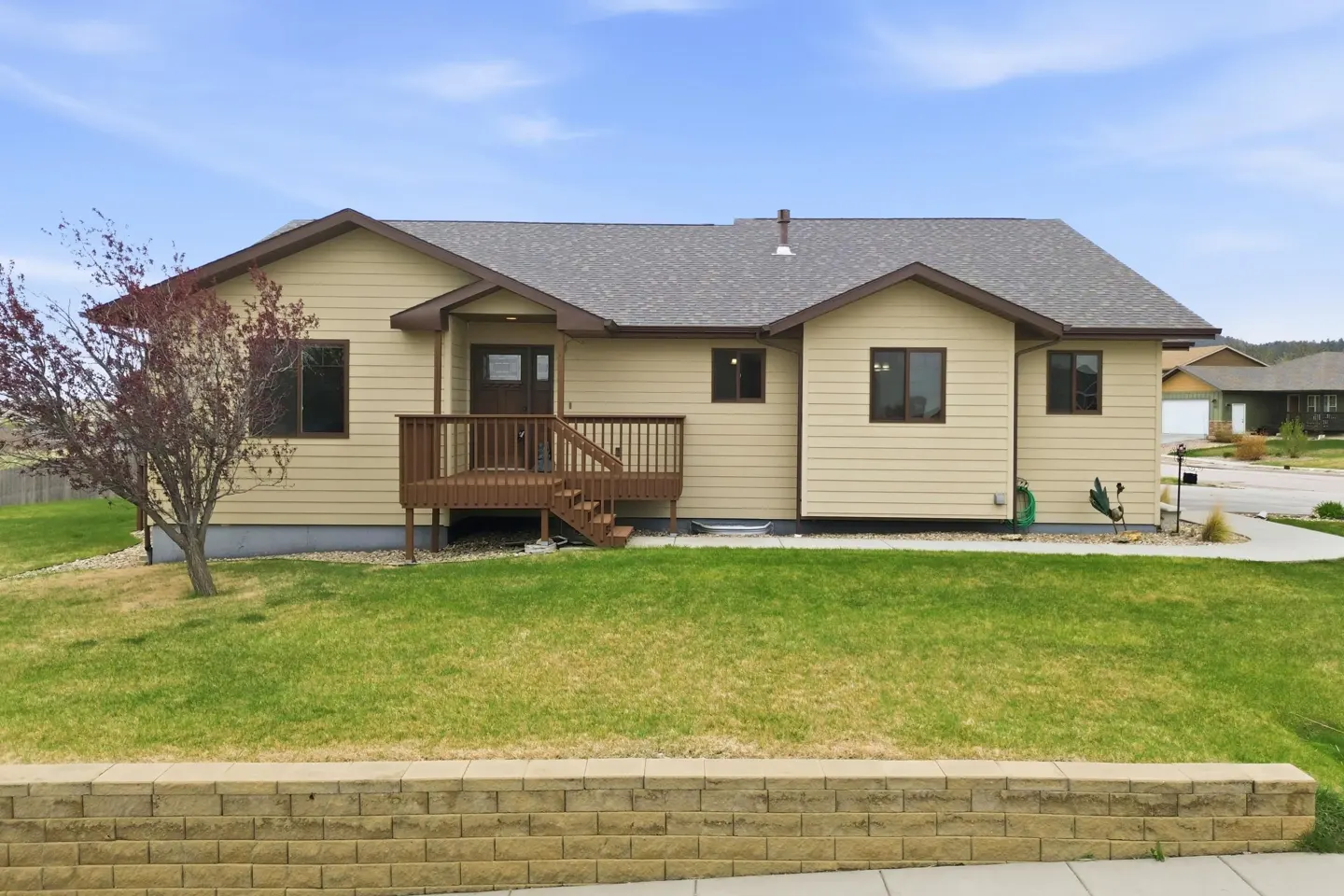 Tan single-story house with a brown deck, gray roof, and green lawn under a blue sky.