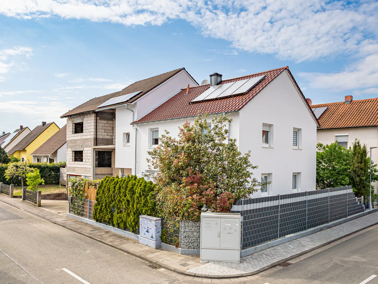 A white two-story house with solar panels on a red roof, next to a house under construction.