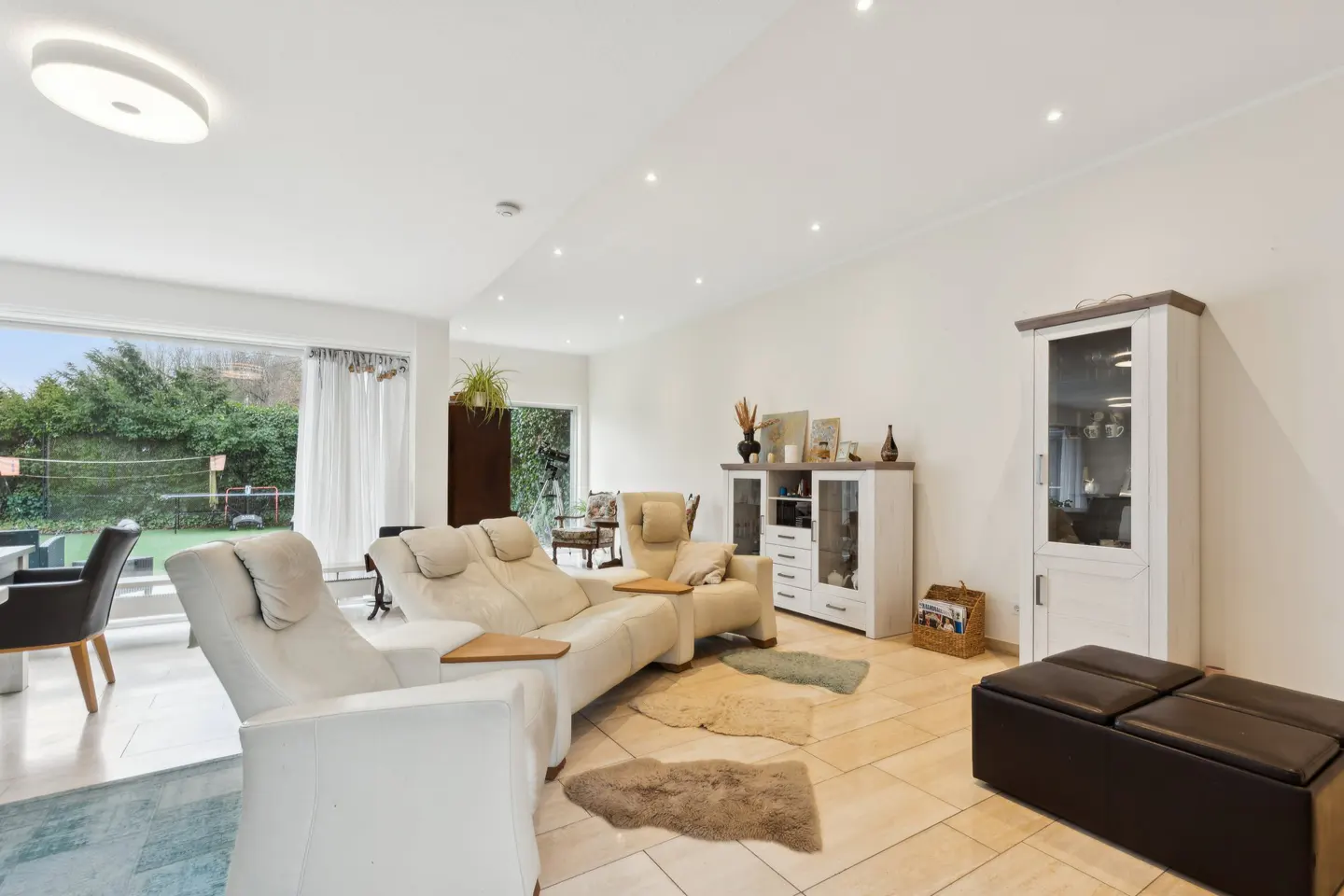 Bright living room with white walls, tile floors, and large windows overlooking a green yard. Beige reclining sofas and white cabinets furnish the space.