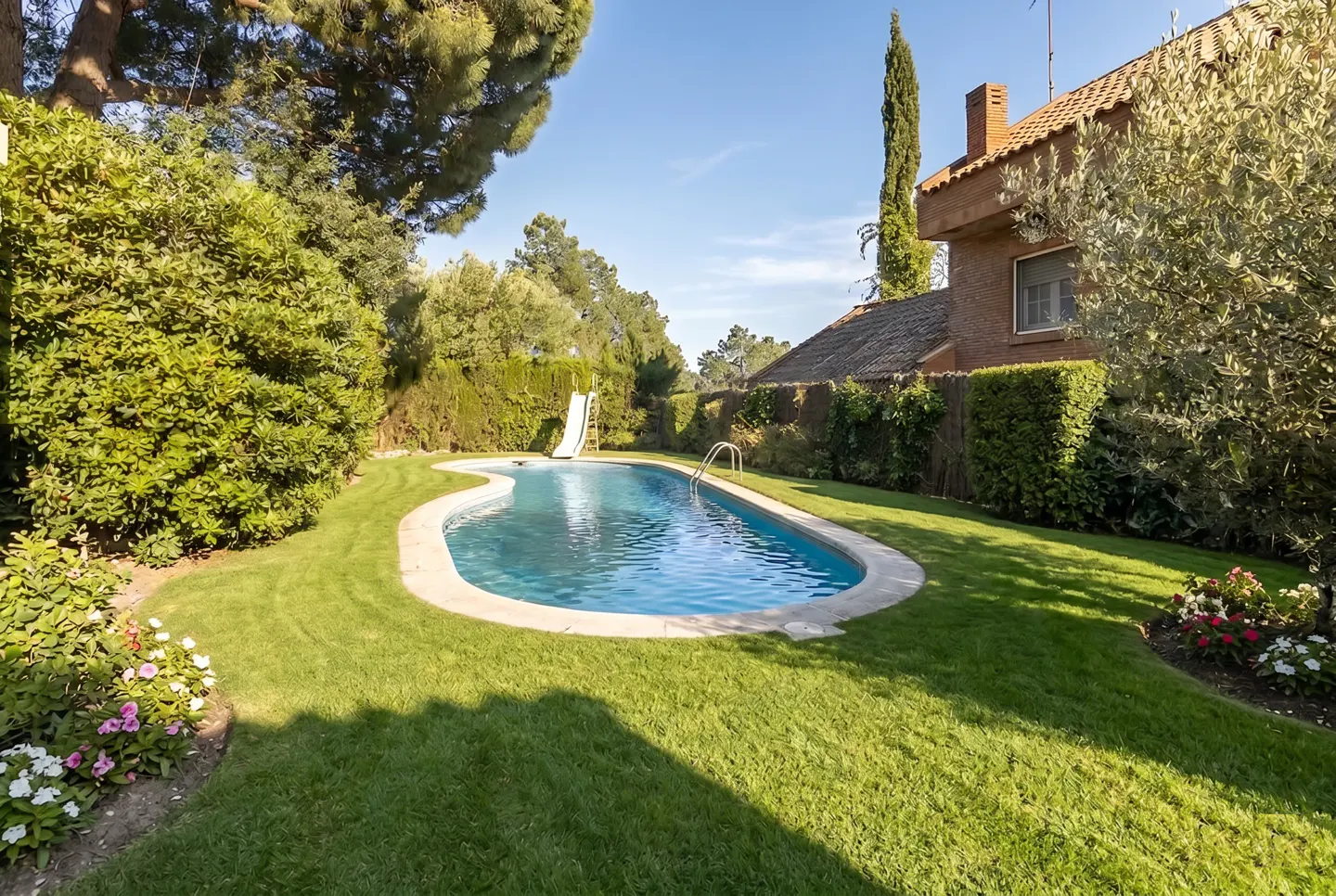 Backyard view of a kidney-shaped pool with a slide, surrounded by green grass, trees, and a brick house.