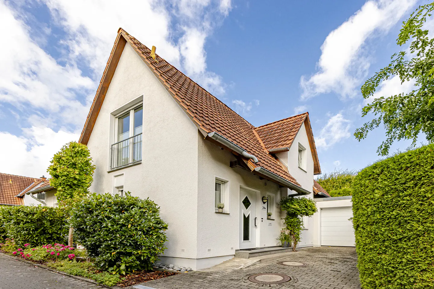 A white, two-story house with a red tile roof, green bushes, and a paved driveway under a blue sky.