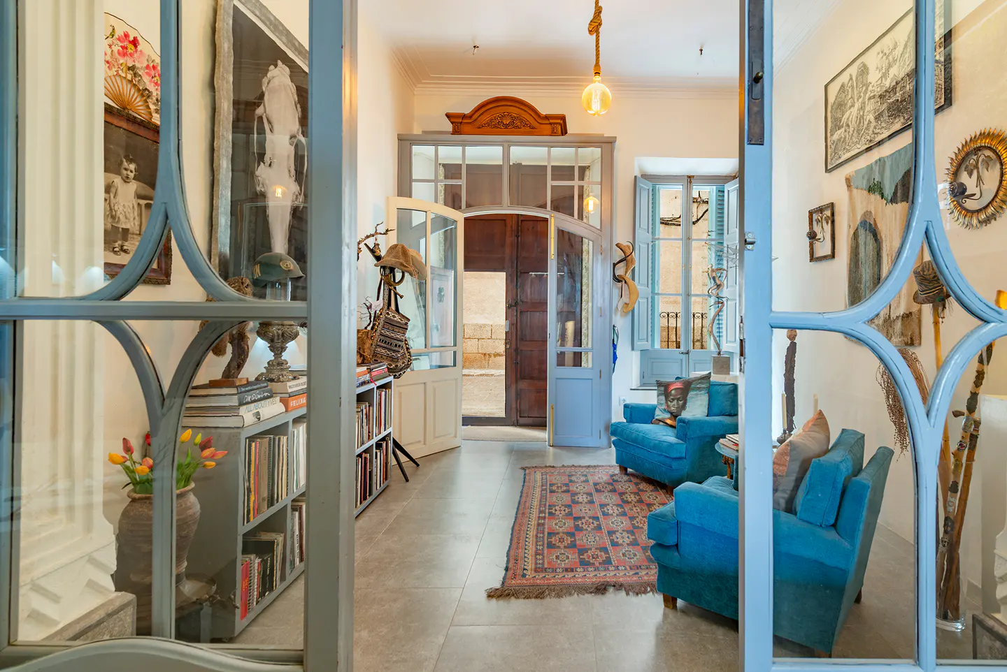 View through ornate blue doors into a bright living room with blue armchairs, art, and an open doorway to the outside.