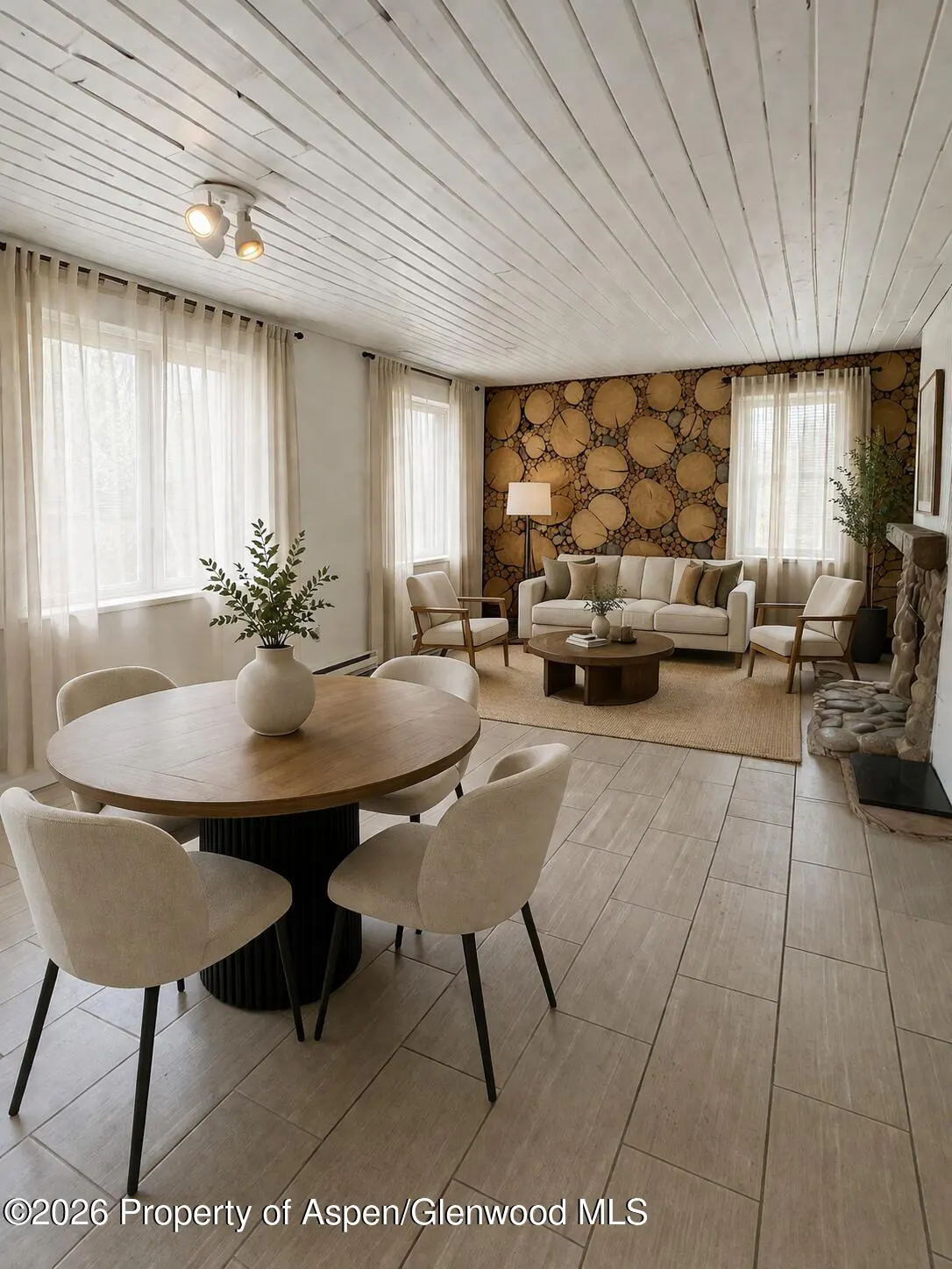 Open-concept living and dining area with a round wood table, beige chairs, and a log-covered accent wall. Cream sofa and stone fireplace.