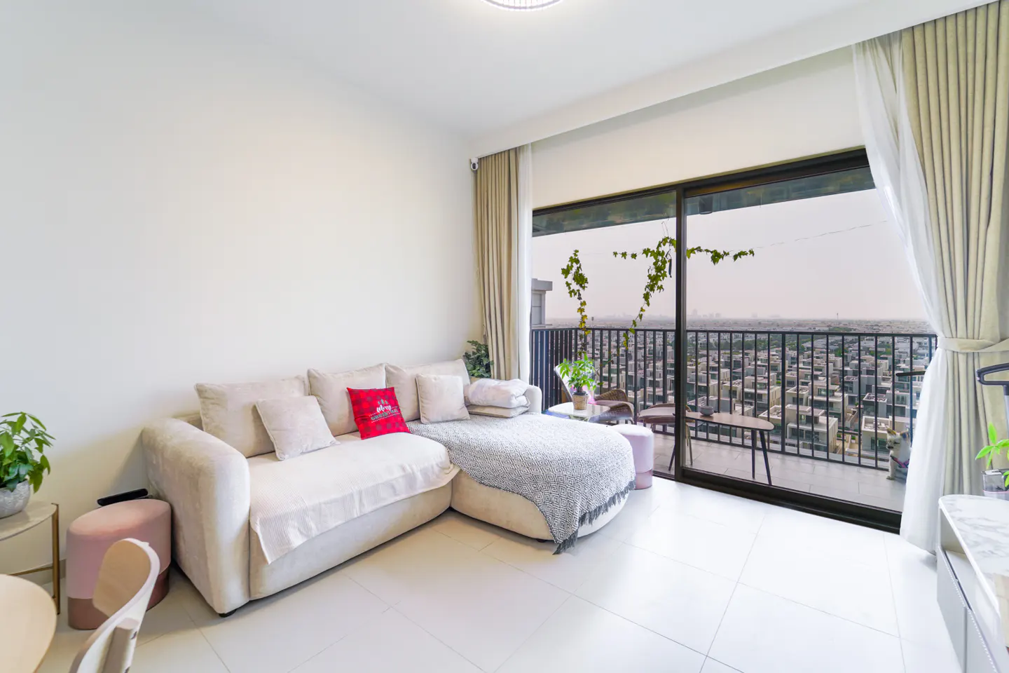 Bright living room with a white sectional sofa, pink accents, and a balcony overlooking a cityscape. Neutral curtains frame the large sliding glass doors.