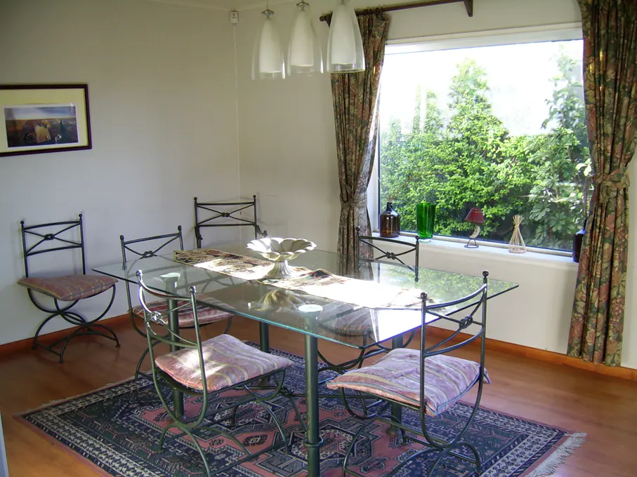 Dining room with glass table, metal chairs with patterned cushions, and a patterned rug. A window with floral curtains overlooks green trees.