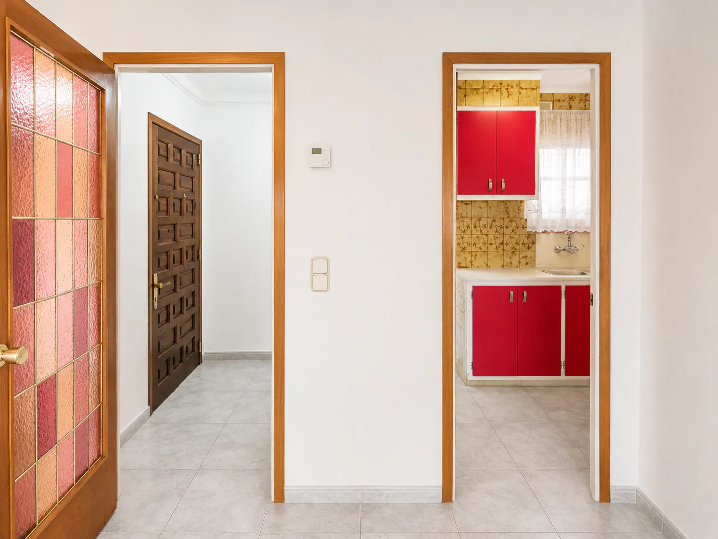 Interior view of a home with three doorways. One has a stained glass door, another leads to a dark wood door, and the third reveals a kitchen with red cabinets.
