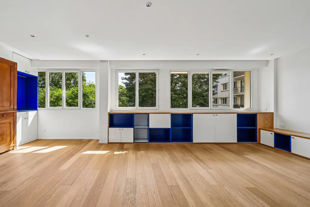 Bright, empty room with wood floors, white walls, and built-in blue and white cabinets under windows with green tree views.