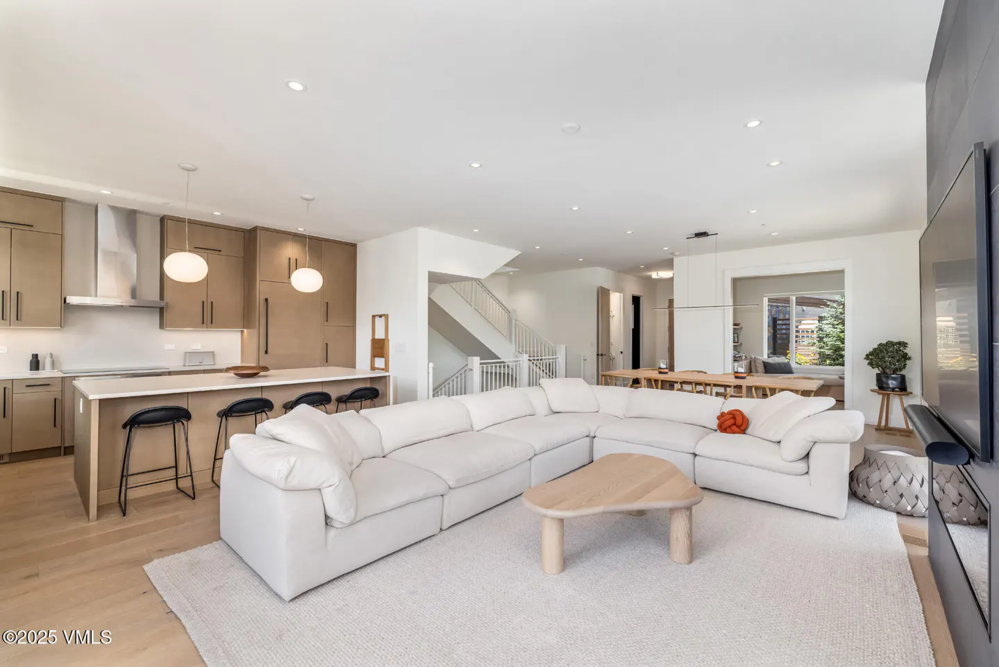 Open-concept living space with a white sectional sofa, wood coffee table, and kitchen island with stools. Neutral tones and natural light.