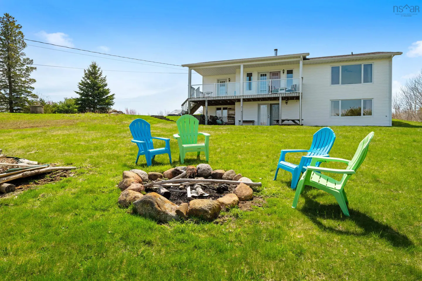Lawn with fire pit surrounded by blue and green Adirondack chairs. A white two-story house is in the background.