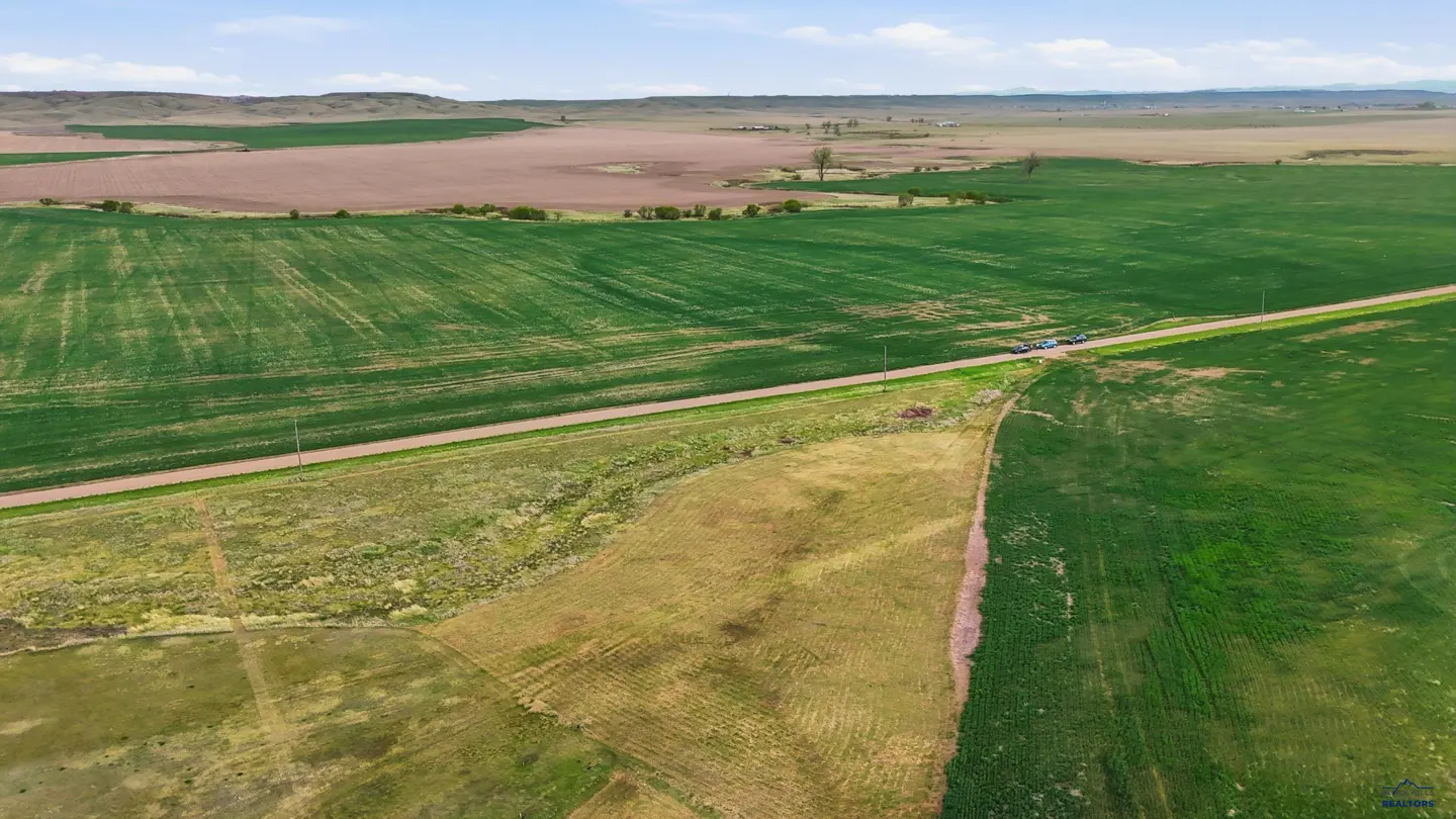 Aerial view of a rural landscape with green and brown fields, a dirt road, and parked cars.
