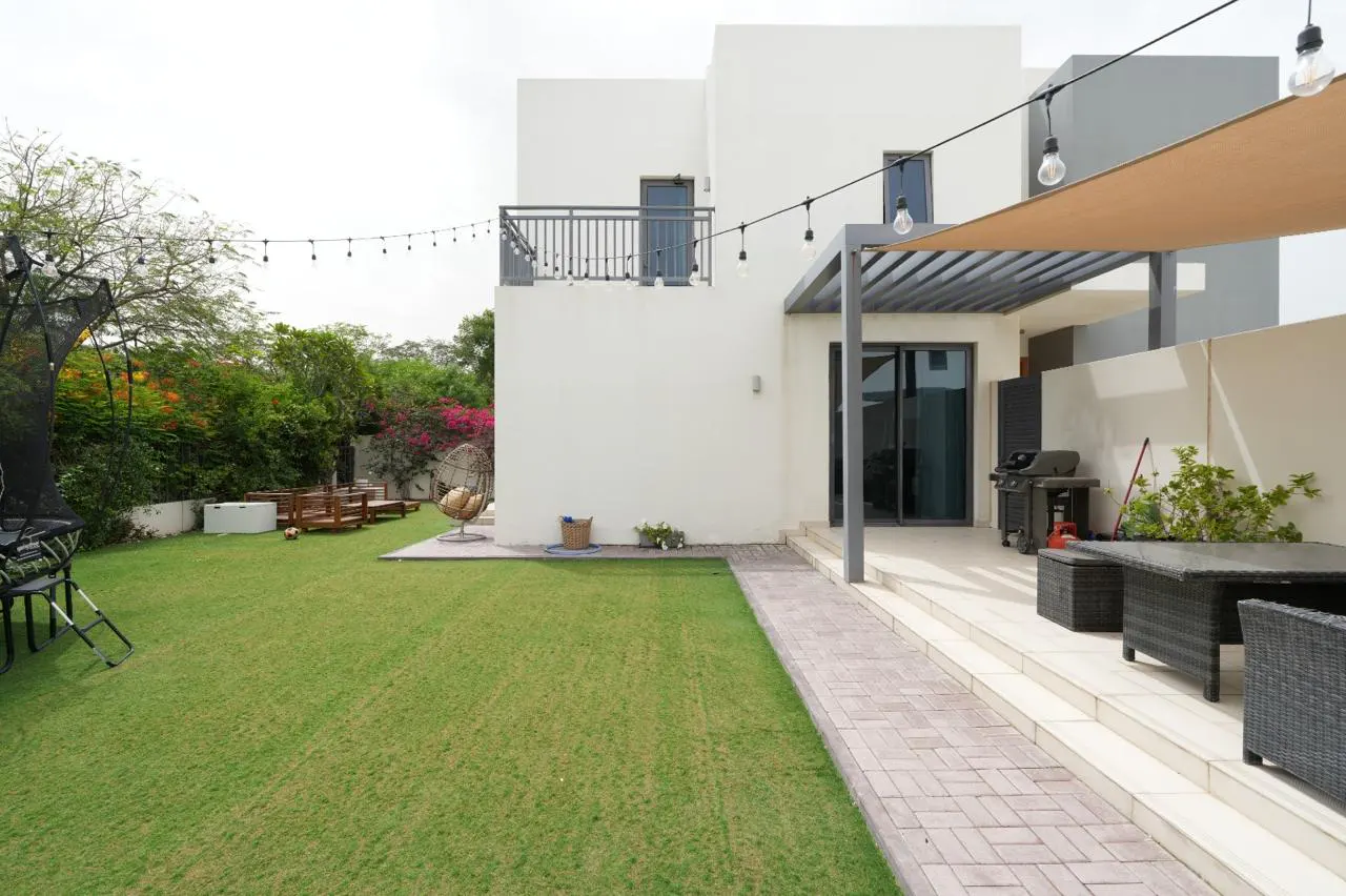 A modern two-story house with a large green lawn, patio, and outdoor furniture. String lights hang above the patio.