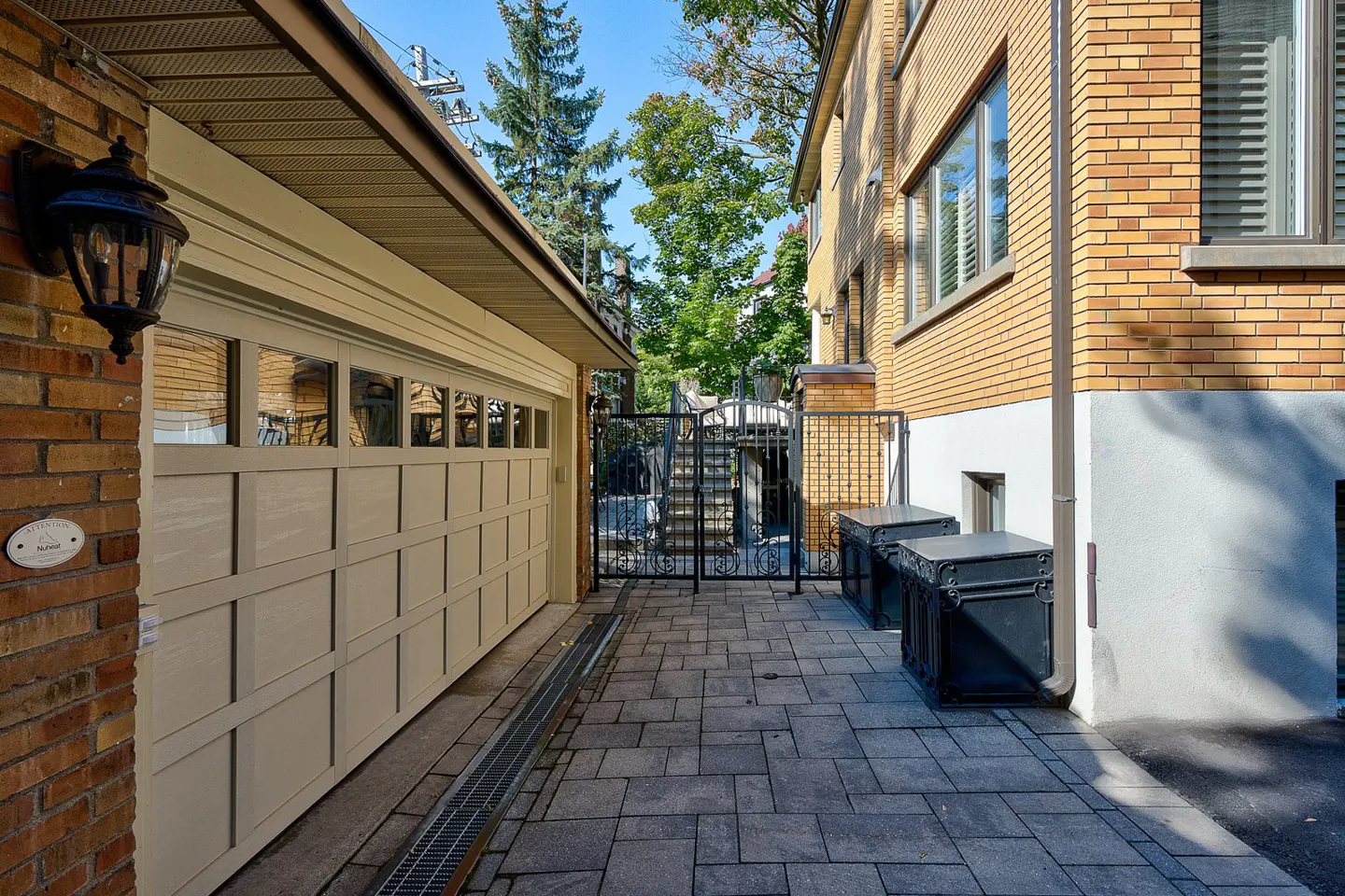 A brick walkway between a beige garage and a brick house leads to a black metal gate.
