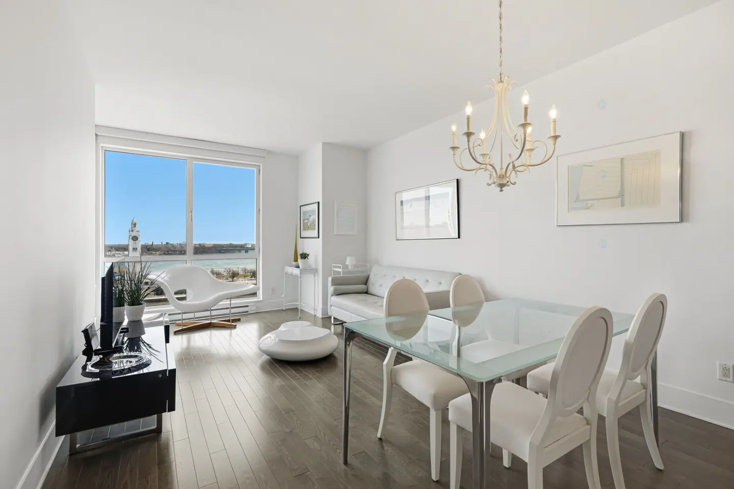 Bright, modern apartment interior with white walls, dark wood floors, and a view of the water. A glass table with white chairs sits under a chandelier.