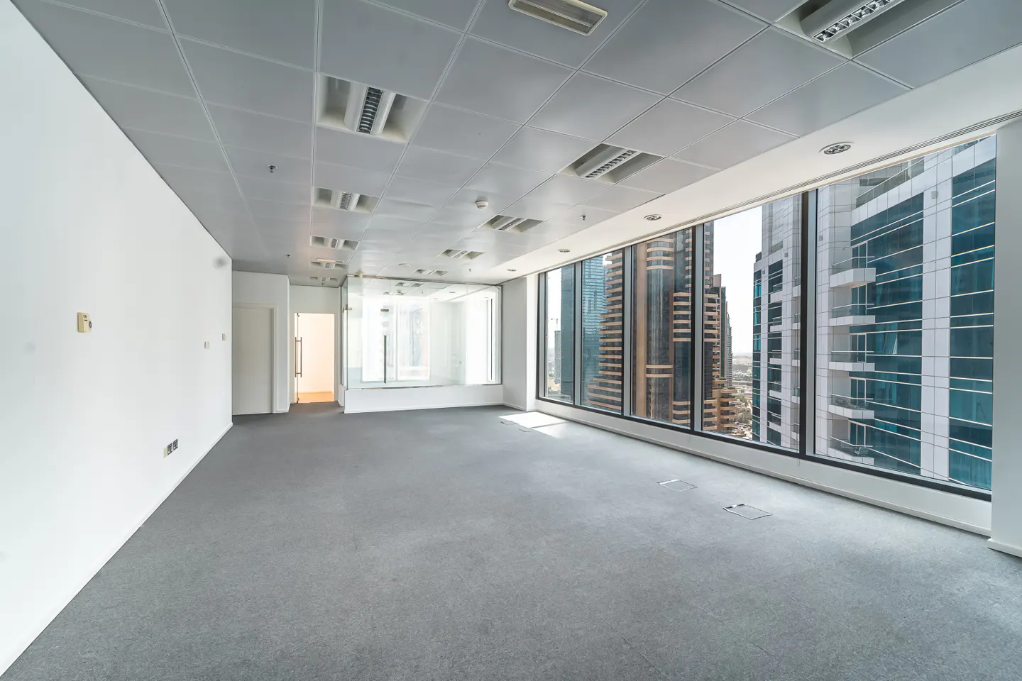 Empty office space with gray carpet, white walls, and a large window showing city buildings.