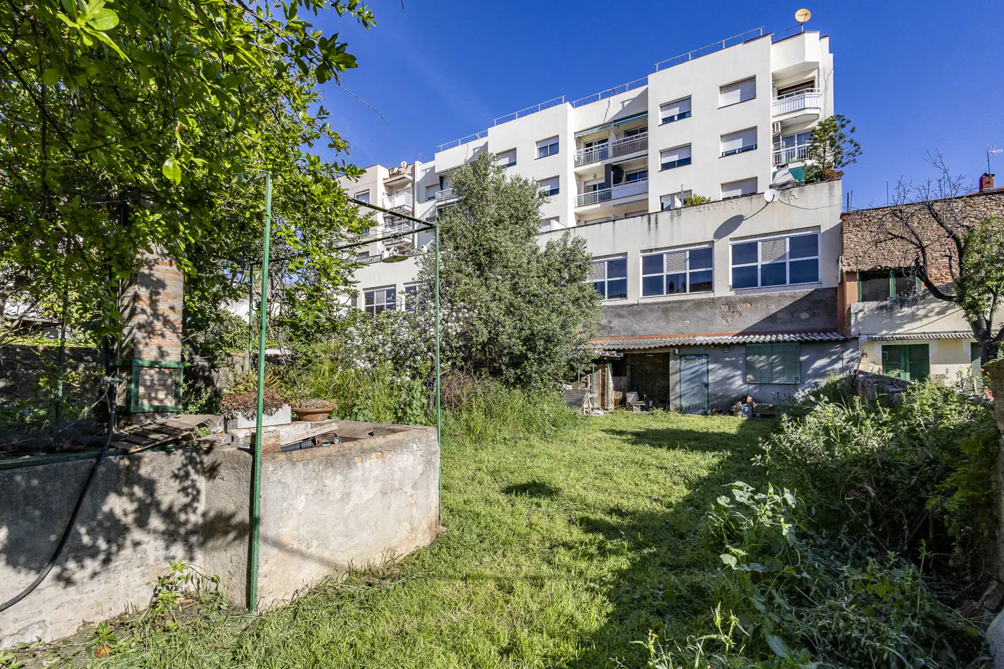 A backyard with a concrete well, brick chimney, and green grass leads to a white building under a blue sky.