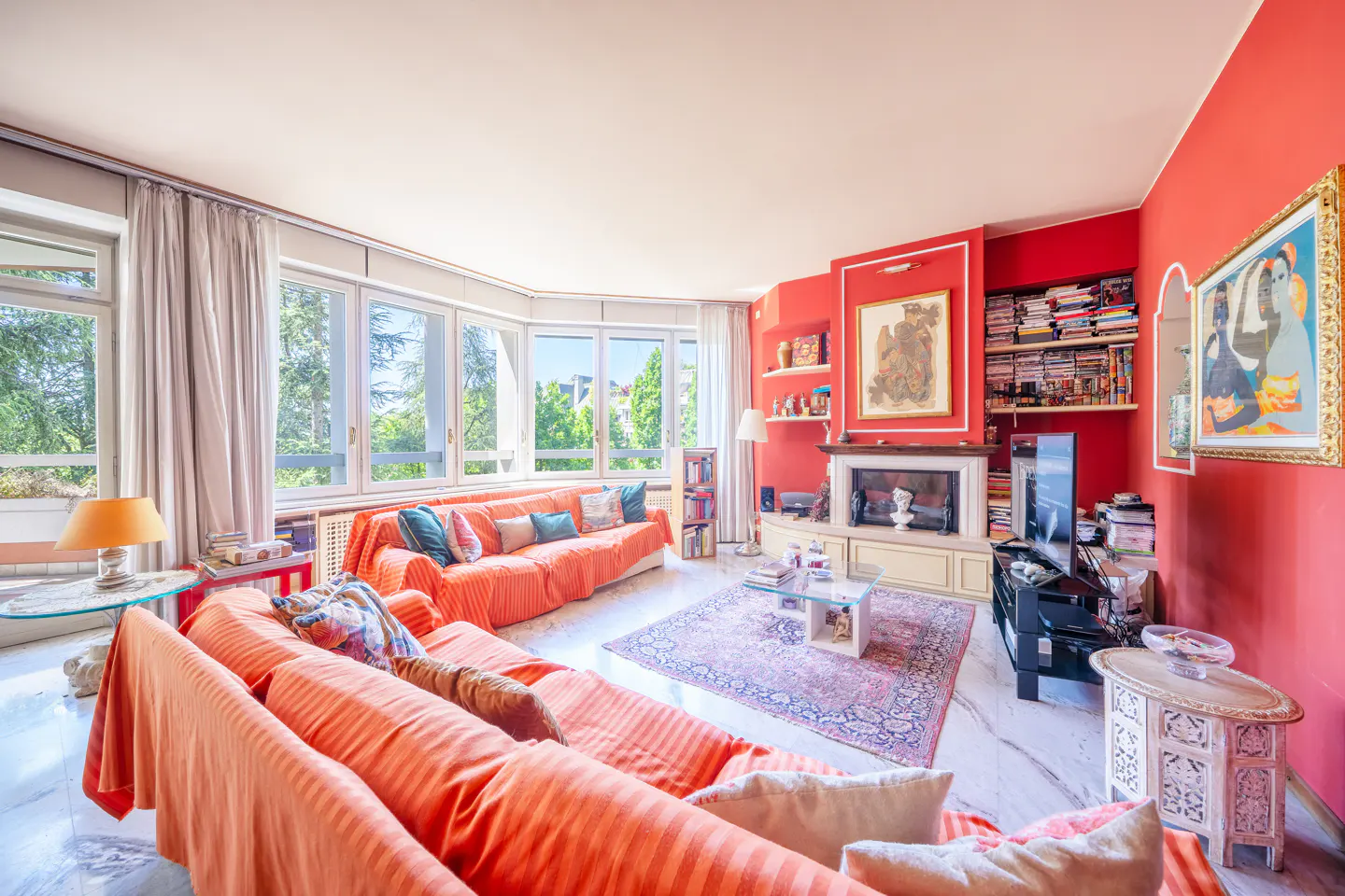 Living room with red walls, orange sofas, marble floors, fireplace, and large windows with green trees outside.