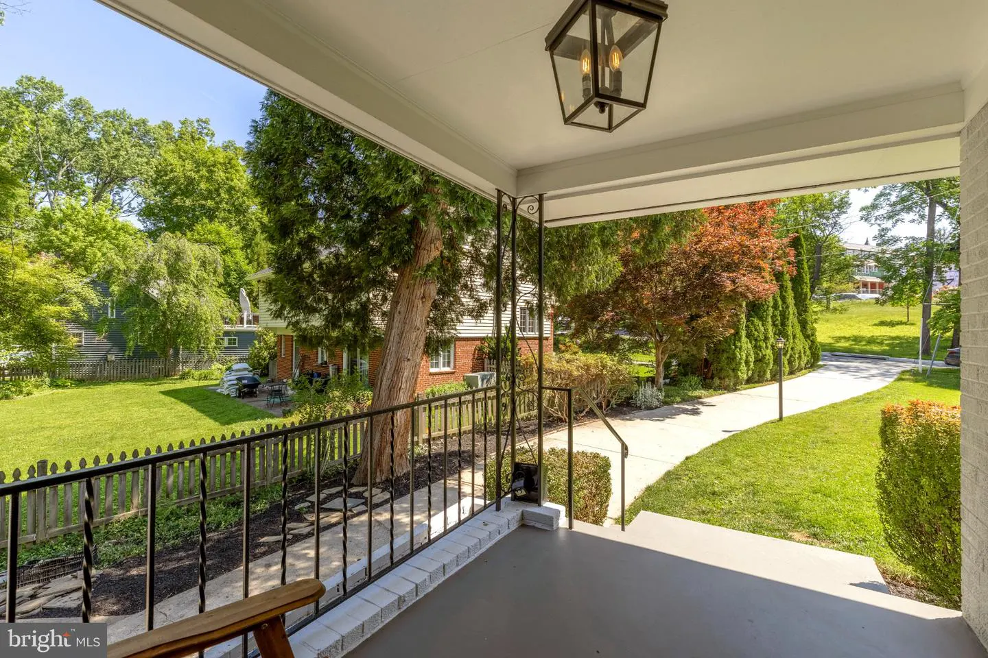 View from a porch with black railing, looking out to a green lawn, trees, and a driveway. A black lantern hangs from the porch ceiling.