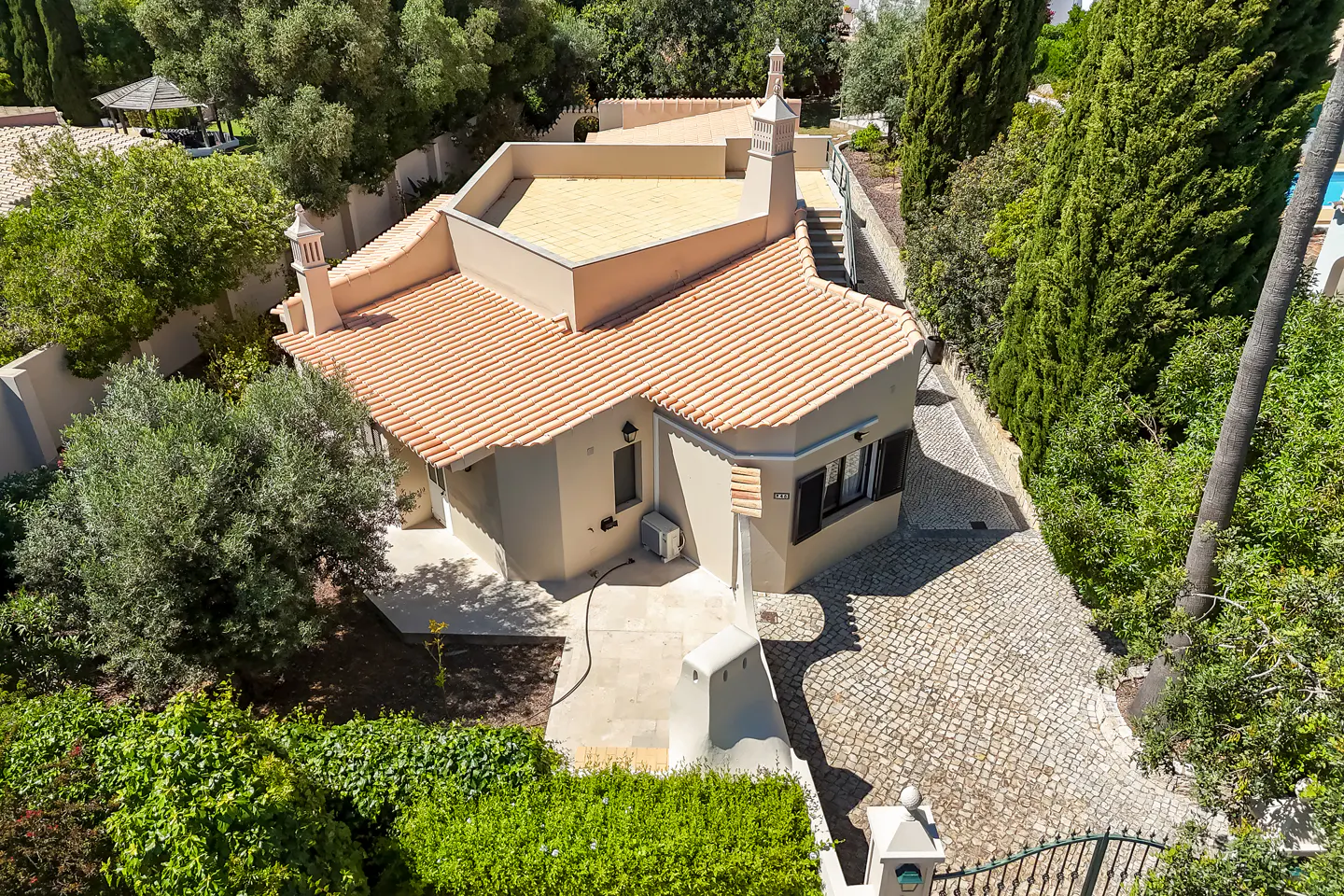 Aerial view of a beige single-story house with a red tile roof, surrounded by lush green trees and a stone driveway.