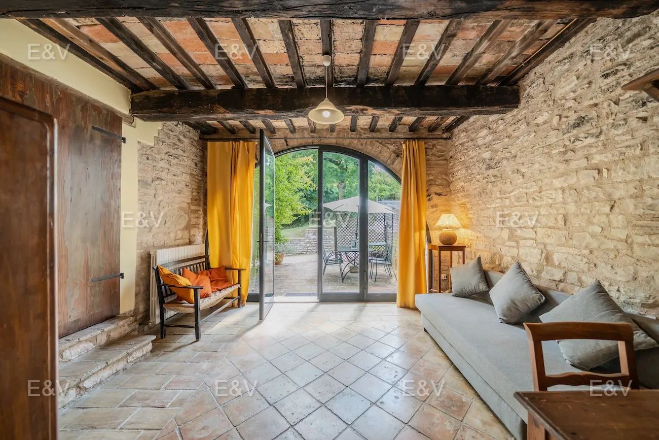 Rustic living room with stone walls, exposed beam ceiling, and terracotta tile floor. French doors open to a patio with outdoor furniture.