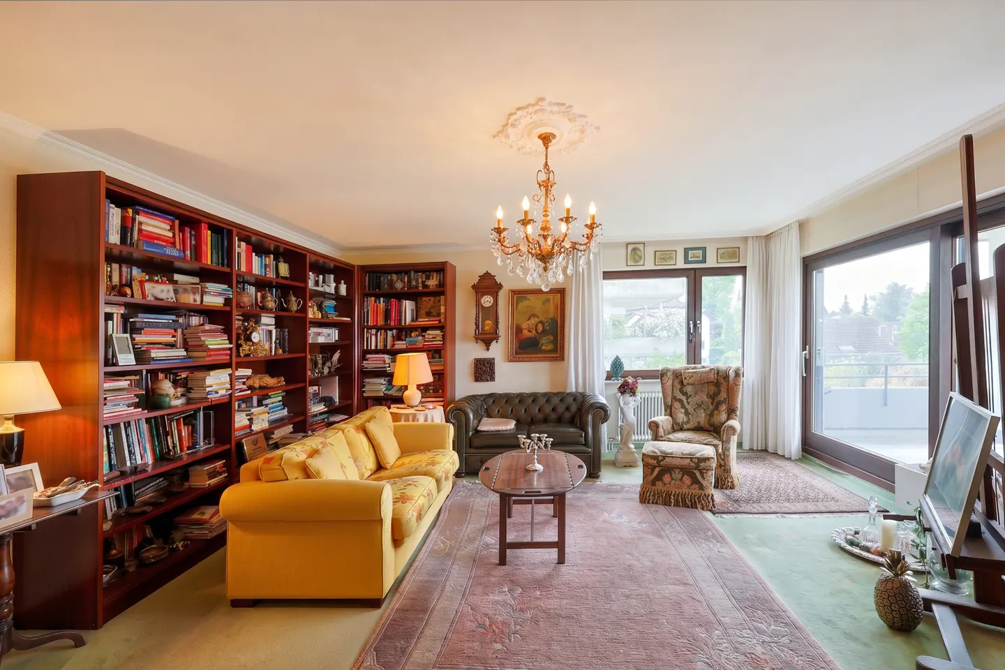 Cozy living room with a yellow sofa, dark leather loveseat, patterned armchair, large bookshelves, and a crystal chandelier.