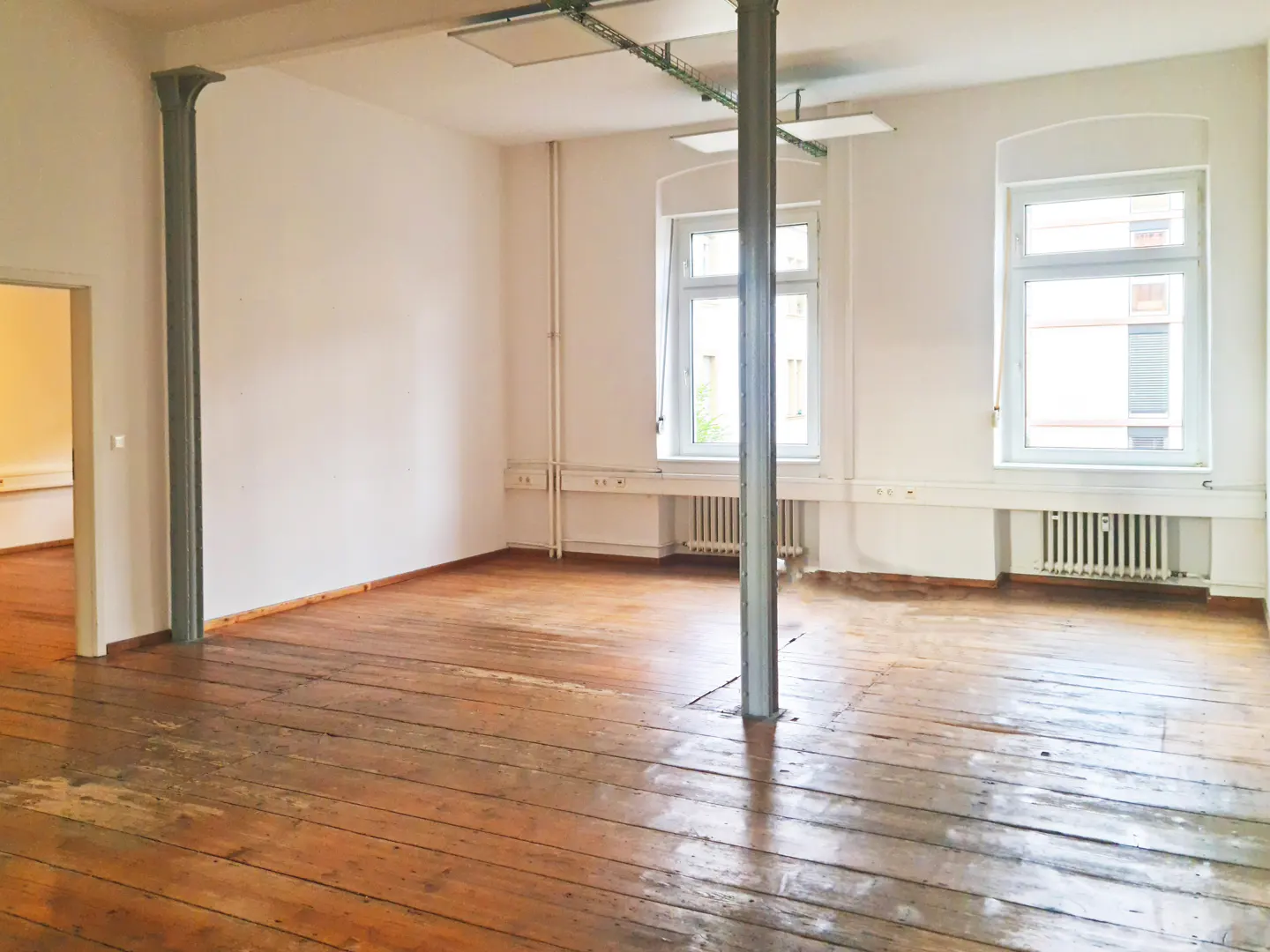 Empty room with white walls, wooden floors, two large windows, and industrial-style metal support pillars.
