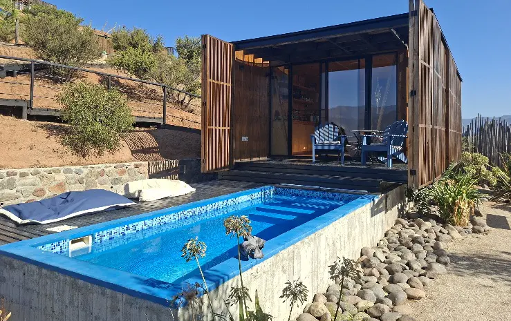A modern wood cabin with a blue pool, two blue chairs, and a table on the patio. The cabin is on a hillside with trees and a blue sky.