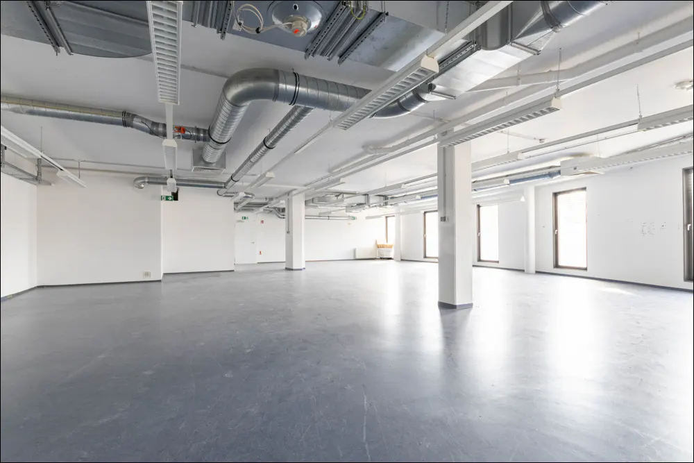 Empty office space with gray floor, white walls, and exposed ceiling with ductwork and lighting. Windows line the far wall.