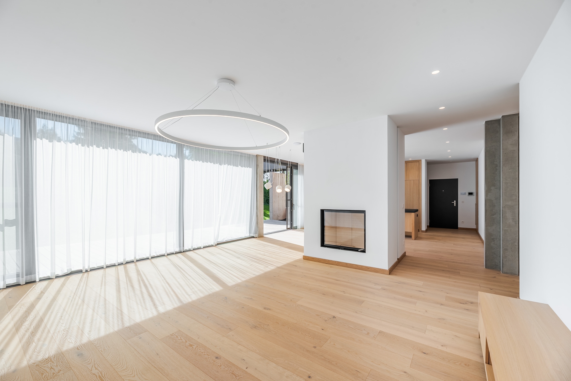 Bright, modern living room with light wood floors, white walls, and a fireplace. Large windows with sheer curtains let in sunlight. A circular light fixture hangs from the ceiling.