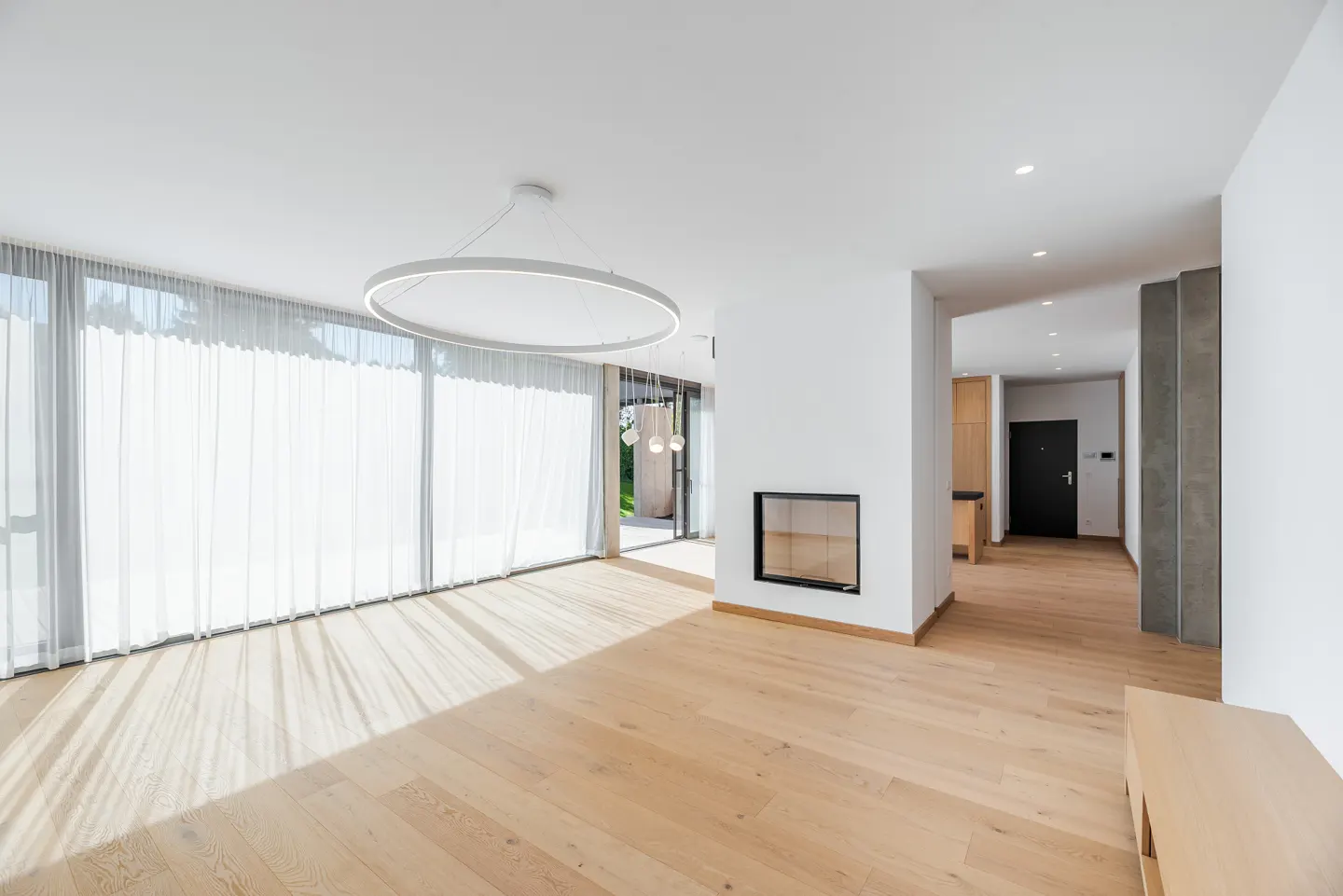 Bright, modern living room with light wood floors, white walls, and a fireplace. Large windows with sheer curtains let in sunlight. A circular light fixture hangs from the ceiling.
